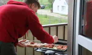 Mężczyzna w czerwonej bluzie przygotowuje mięso i kiełbaski na grillu na balkonie. Idealne miejsce, gdzie można grillować.