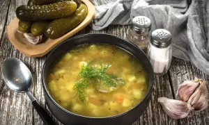 Miseczka zupy ogórkowej, obok ogórki kiszone, czosnek i przyprawy. Idealna do doprawienia, by uzyskać najlepszy smak.