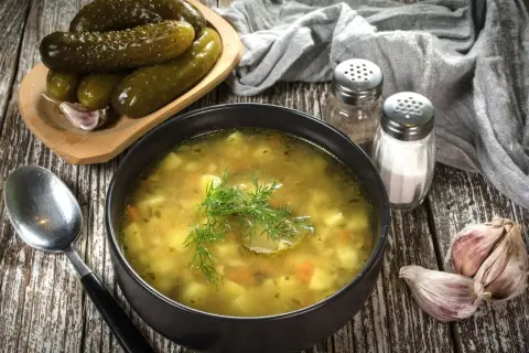 Miseczka zupy ogórkowej, obok ogórki kiszone, czosnek i przyprawy. Idealna do doprawienia, by uzyskać najlepszy smak.
