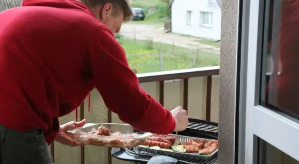 Mężczyzna w czerwonej bluzie przygotowuje mięso i kiełbaski na grillu na balkonie. Idealne miejsce, gdzie można grillować.