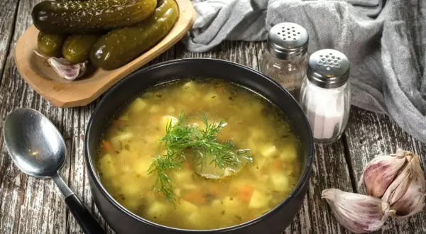 Miseczka zupy ogórkowej, obok ogórki kiszone, czosnek i przyprawy. Idealna do doprawienia, by uzyskać najlepszy smak.