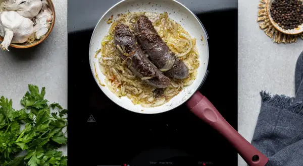 Kaszanka smażona na patelni z cebulą, obok czosnek, pieprz i natka pietruszki.