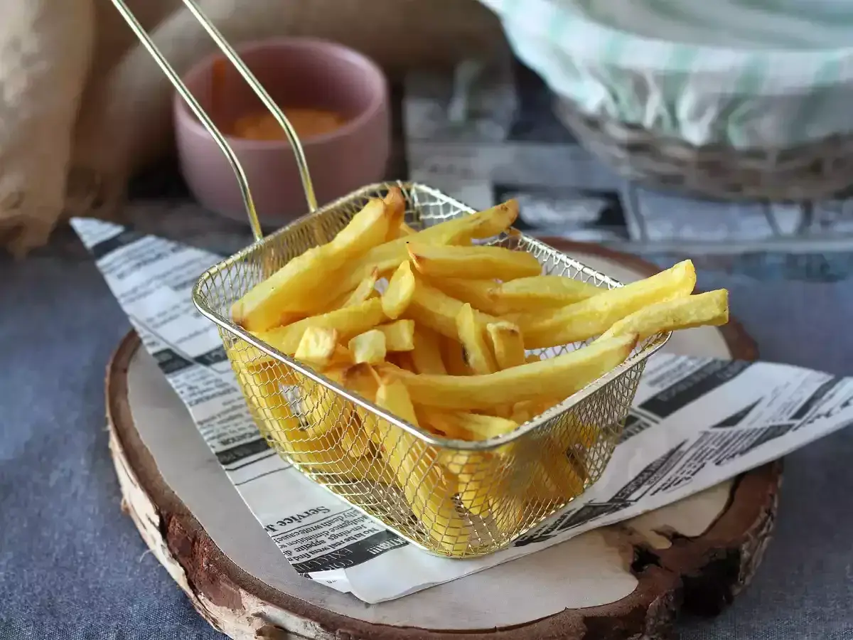 Złota, chrupiąca frytka w metalowym koszyku na tle sosu i koszyka.