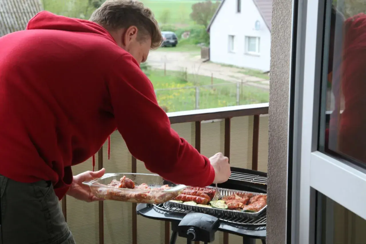 Mężczyzna w czerwonej bluzie przygotowuje mięso i kiełbaski na grillu na balkonie. Idealne miejsce, gdzie można grillować.