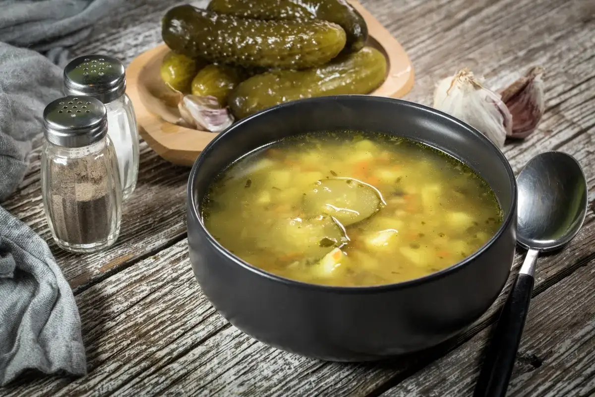 Zupa ogórkowa gotowa do doprawienia. Obok miski z zupą leżą ogórki kiszone, czosnek, solniczki i łyżka.
