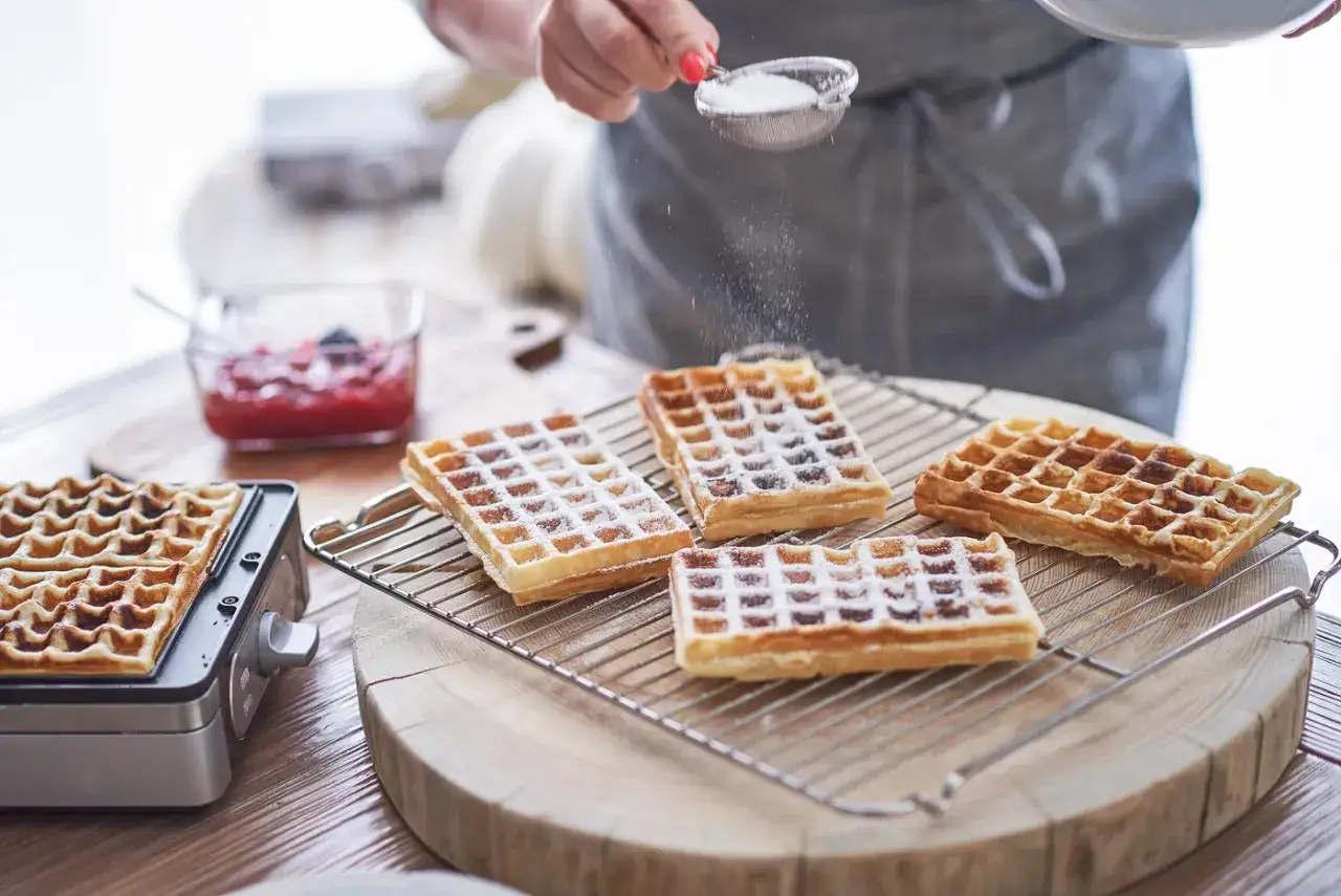 Jak upiec wafle? Na stole świeżo upieczone, złociste wafle posypywane cukrem pudrem. Obok dżem i gofrownica.