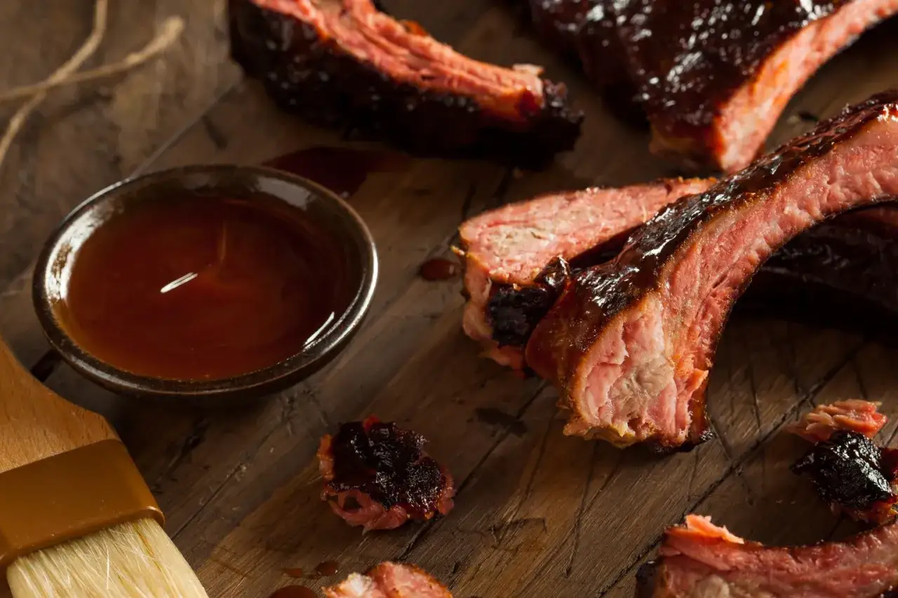 Pyszne, glazurowane żeberka BBQ na drewnianej desce. Obok miseczka z sosem i pędzelek.