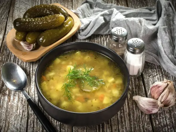 Miseczka zupy ogórkowej, obok ogórki kiszone, czosnek i przyprawy. Idealna do doprawienia, by uzyskać najlepszy smak.